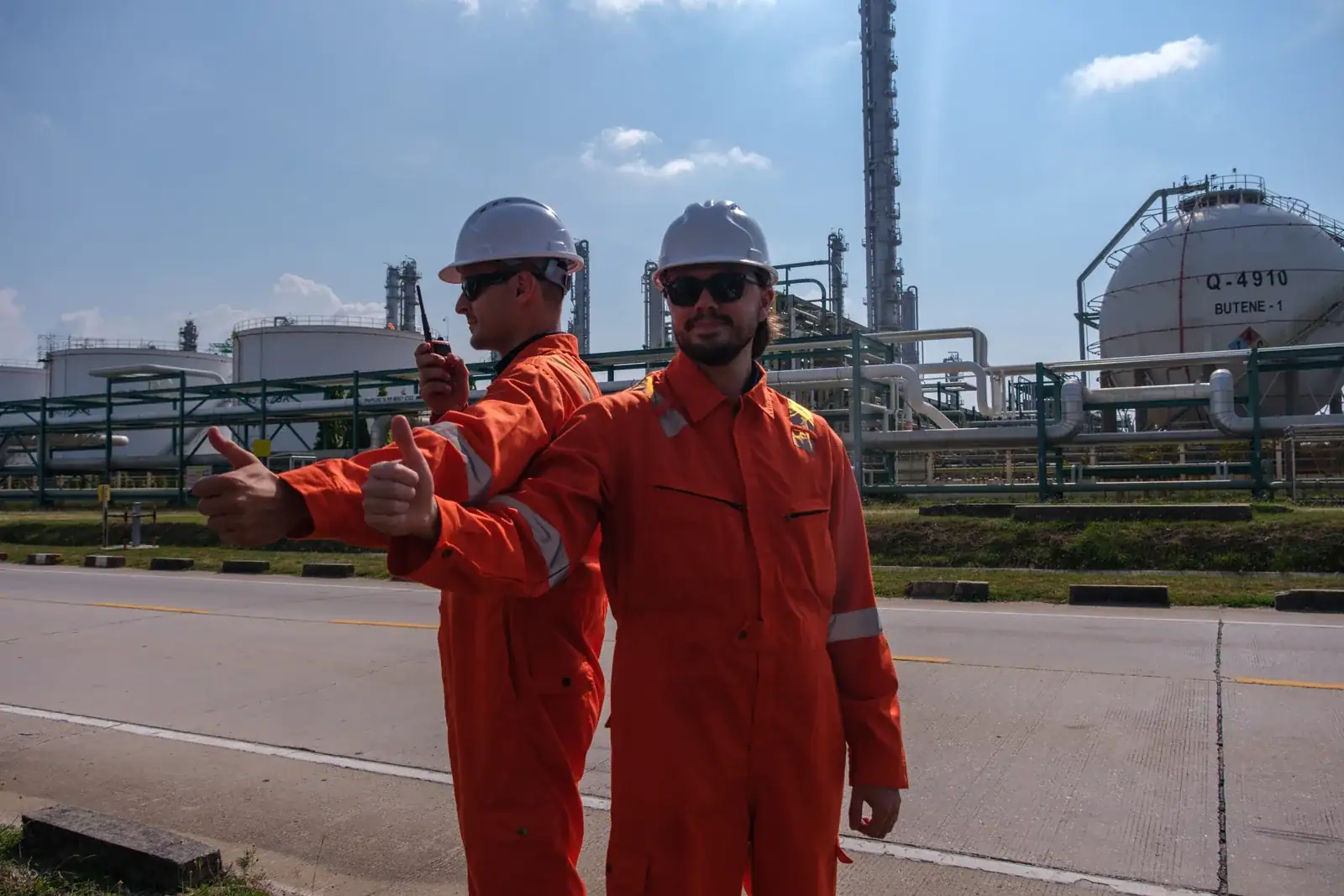 Two engineers in orange coveralls beside industrial storage tanks.