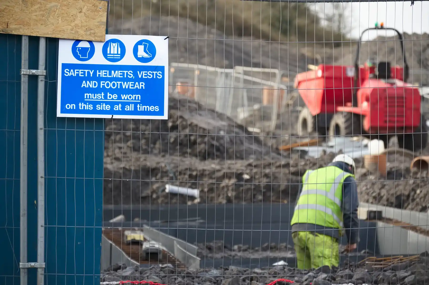 Construction site safety sign on a fence beside a worker and a site dumper.