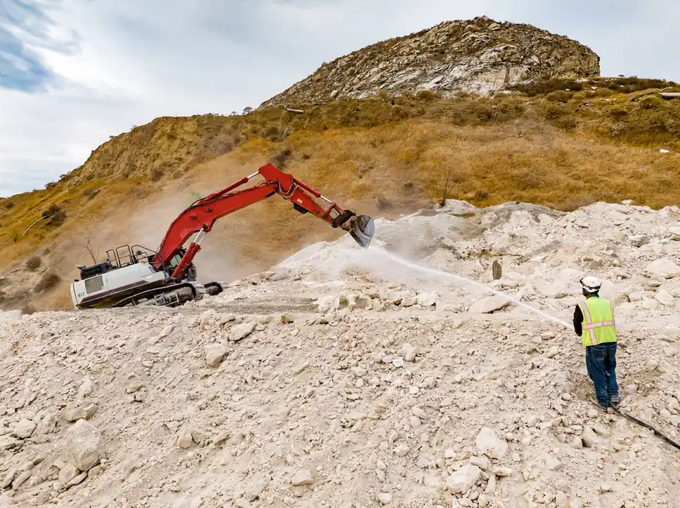 Water being sprayed to suppress dust while an excavator breaks rock.