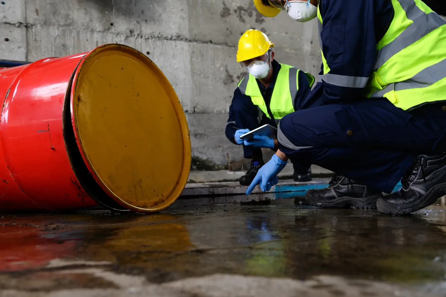 Workers in respirators inspecting a spill beside a fallen drum.