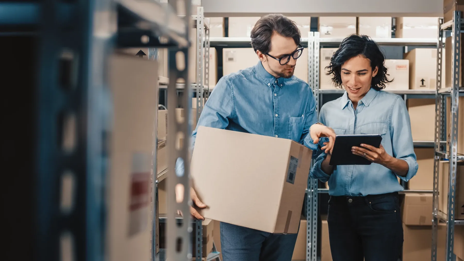 Two warehouse staff checking a tablet while one holds a cardboard box.