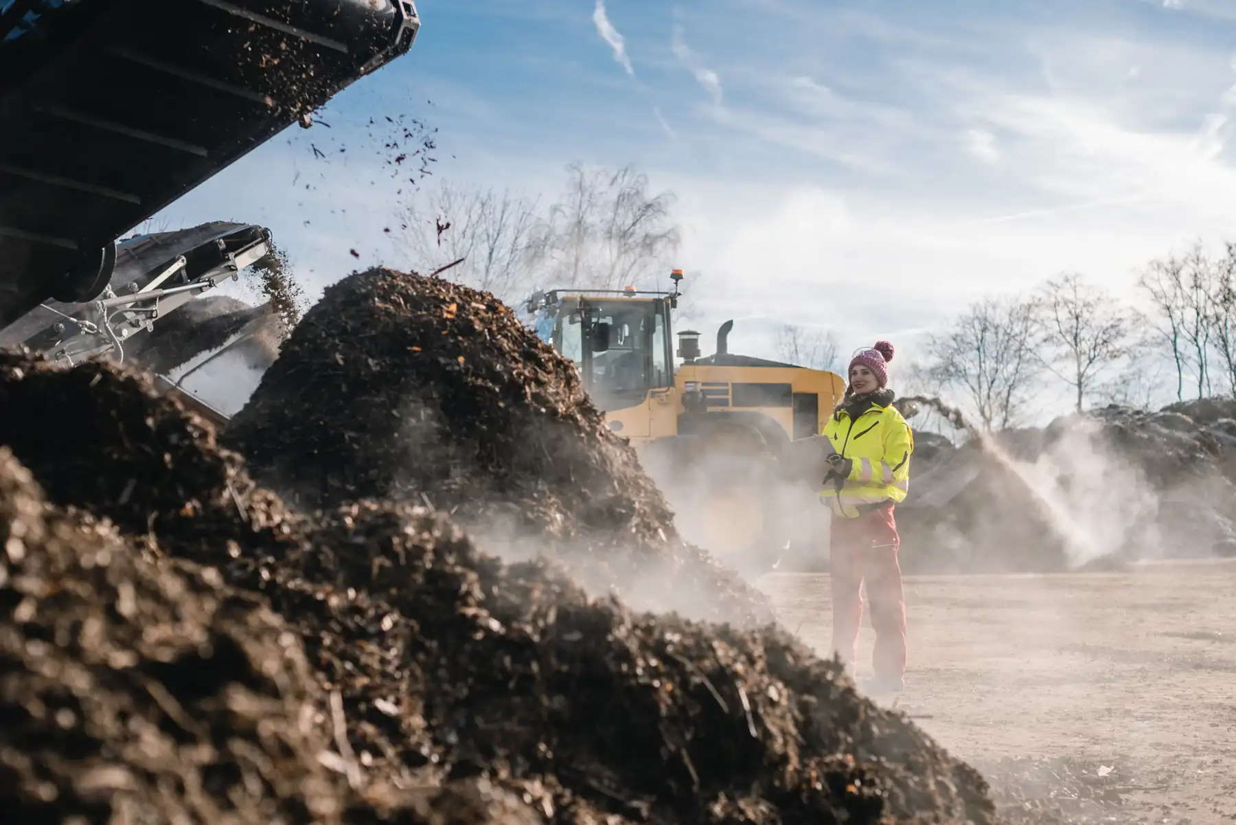 Worker with a clipboard beside a loader and compost heaps at a processing site.