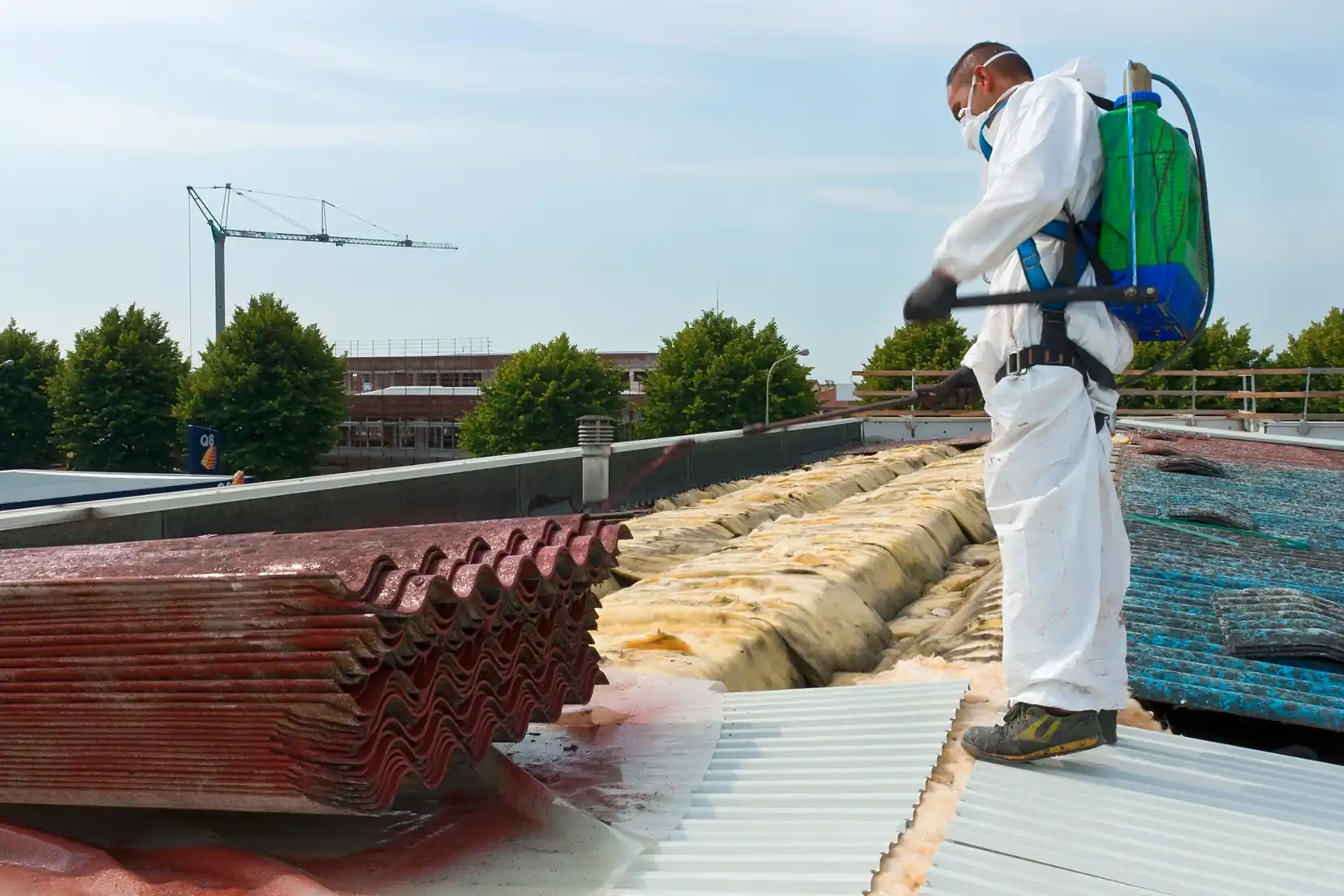 Worker in a protective suit and sprayer beside removed roofing sheets.